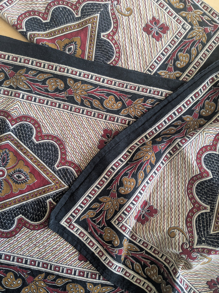 Overlapping pieces of Indian block printed fabric, in rich black, deep red, ochre and cream hues, mixing traditional Indian motifs and geometric lines and shapes.