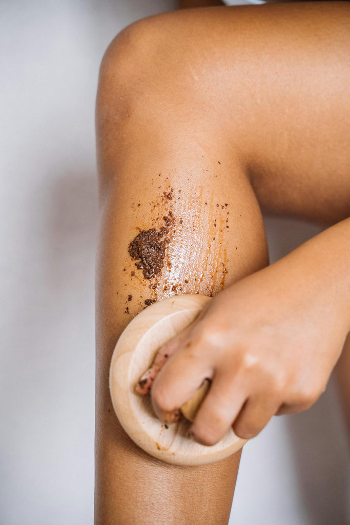 Close-up of a person exfoliating their leg with a natural scrub and a wooden body brush. The rich texture of the scrub and warm skin tone evoke a calming, earthy self-care ritual focused on natural skincare and gentle renewal.