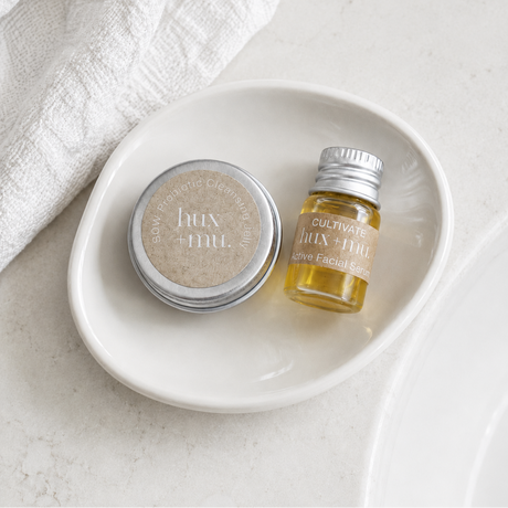 Two discovery sizes of hux and mu products, one round tin and one bottle, on a white plate with a white cloth in the background.
