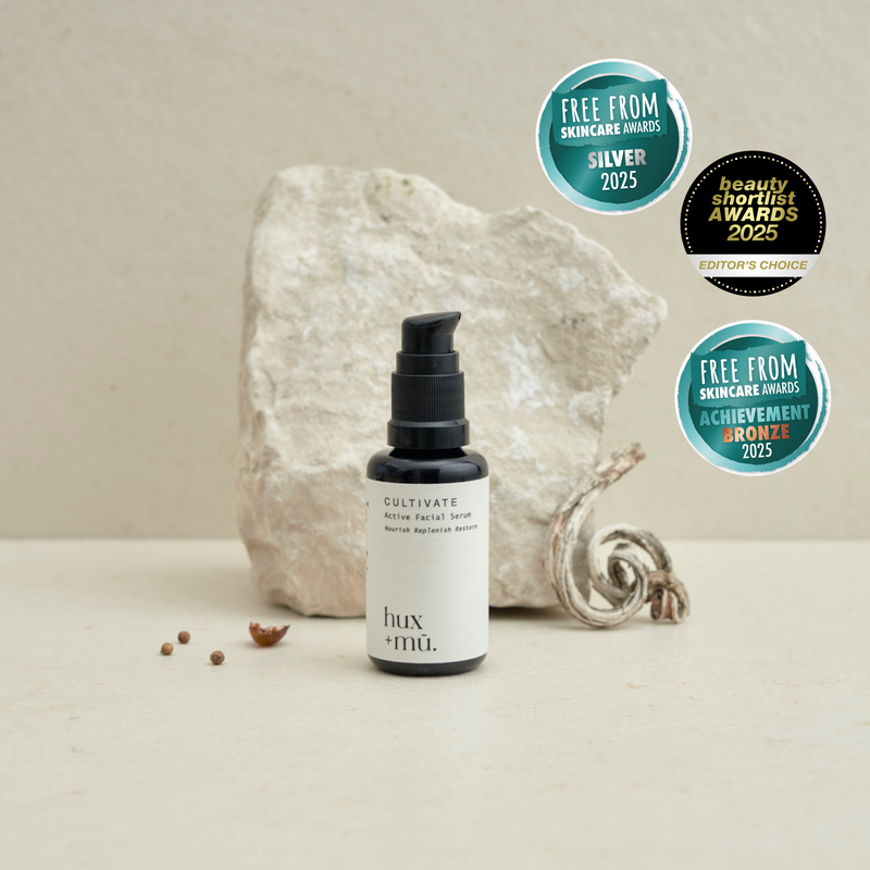 A minimal, elegant product image featuring a dark glass bottle of "Hux + Mü Cultivate Active Facial Serum" with a white label, placed on a light neutral background. The label reads “Nourish Replenish Restore.” Above the product are two teal award badges from the Free From Skincare Awards 2025 — one indicating a Silver award, and the other a Bronze.  There's also a  Beauty Shortlist Editors Choice Award 2025