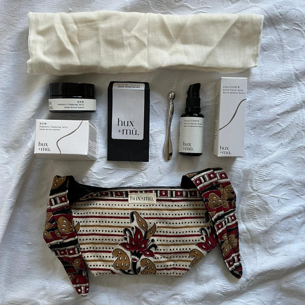 A flat lay style set up of a collection of hux and mu beauty products and a unique block-printed pouch on a white textured cotton surface