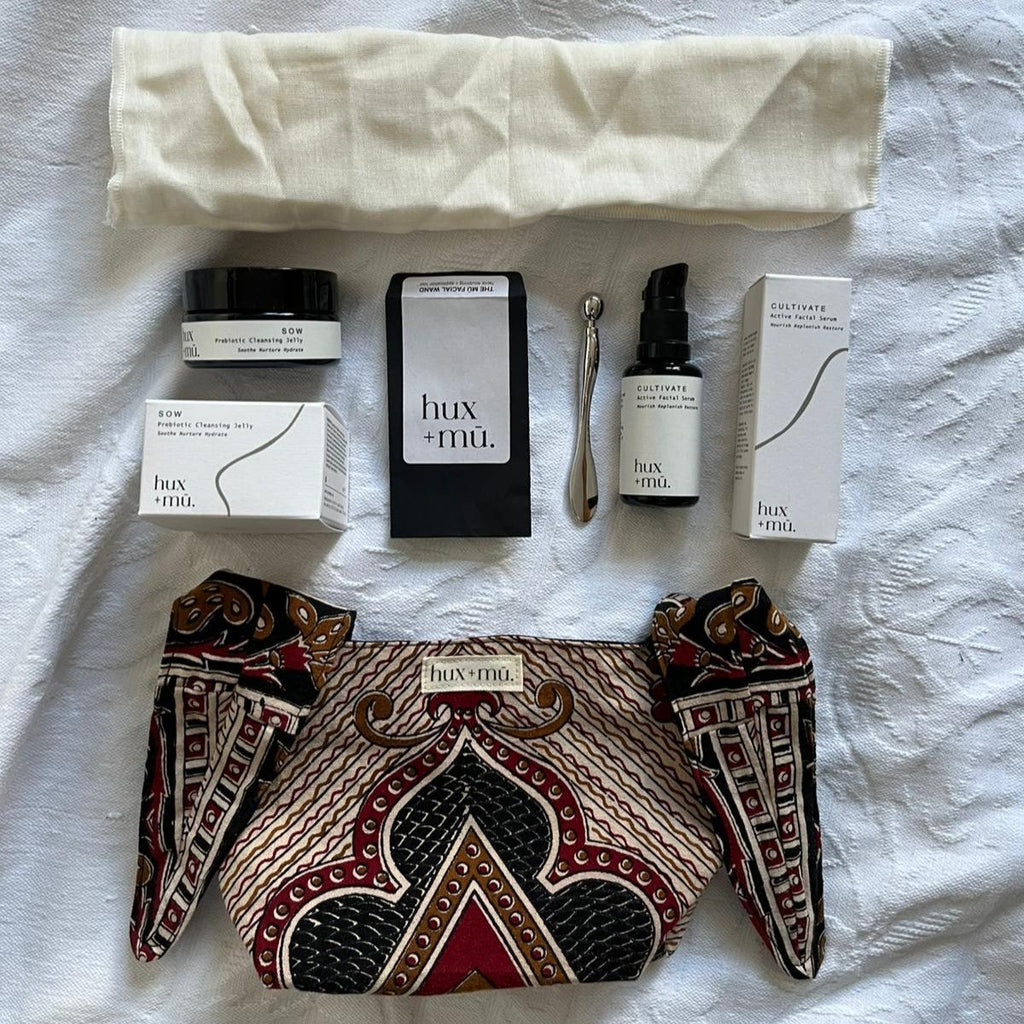 A flat lay style set up of a collection of hux and mu beauty products and a block-printed pouch on a white textured cotton surface