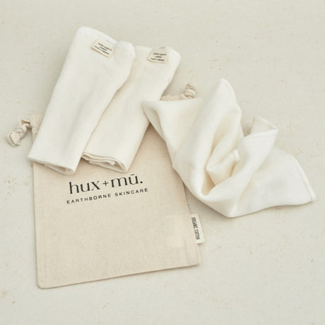 Three organic unbleached muslin face cloths in an organic cotton bag with 'hux+mé' branding on a light background