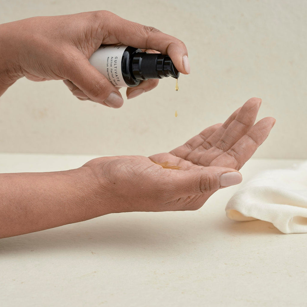 hands pumping drops of hux and mu cultivate active facial serum from bottle in neutral setting with linen face cloth
