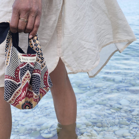 Woman standing in calm clear sea, holding a hux and mu block printed  artisan pouch, in cream, ochre, black and deep red.
