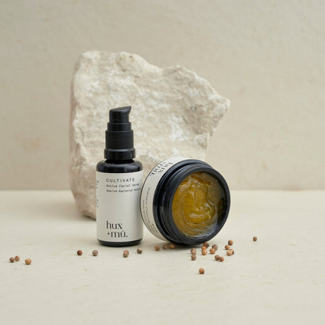 an open jar of hux + mu cleansing jelly  and facial serum bottle in front of stone and amongst coriander seeds